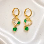 Aros dorados Esmeralda con cristales en forma de corazón verde y piedras brillantes sobre fondo blanco, joyería en Chile inspirada en el lujo cotidiano.