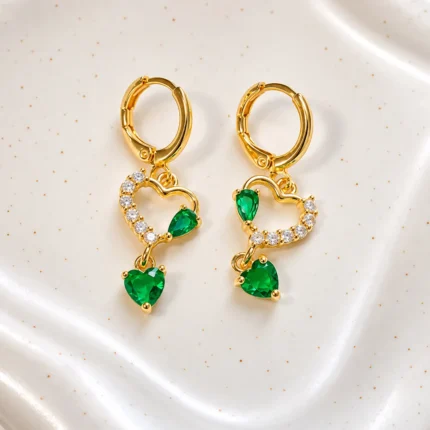 Aros dorados Esmeralda con cristales en forma de corazón verde y piedras brillantes sobre fondo blanco, joyería en Chile inspirada en el lujo cotidiano.