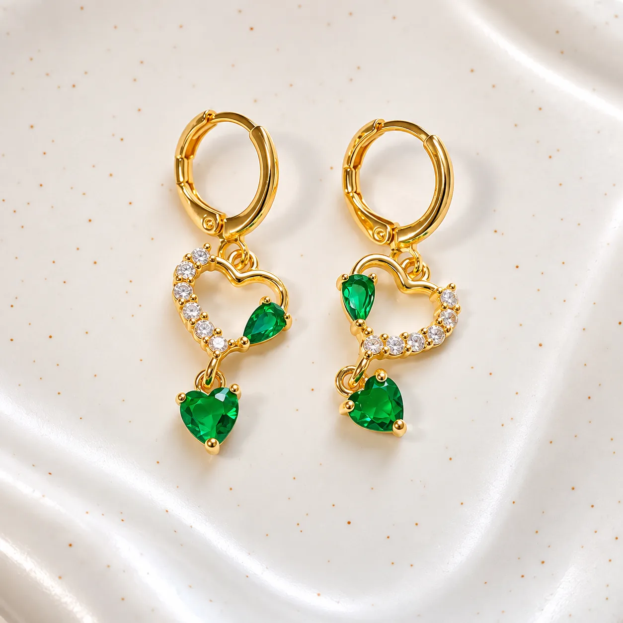 Aros Esmeralda Aros dorados Esmeralda con cristales en forma de corazón verde y piedras brillantes sobre fondo blanco, joyería en Chile inspirada en el lujo cotidiano.