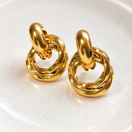 Aros dorados entrelazados Lyra sobre bandeja de cerámica blanca con fondo de satén, joyería en Chile de lujo cotidiano y elegante.